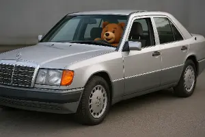 Pohled lehce z boku na stříbrný Mercedes benz w124. Řidič je vysmátý plyšový hnědý medvěd.