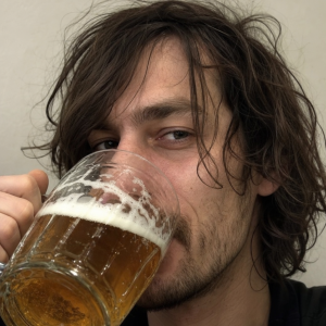 vlasatý muž hubené postavy, pouze obličej, pije pivo z půllitru