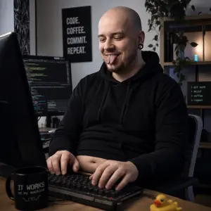 holohlavý ajťák spokojeně píše do klávesnice. Pearcing v obočí, split jazyka, který mu kouká z pusy, trochu při těle.
